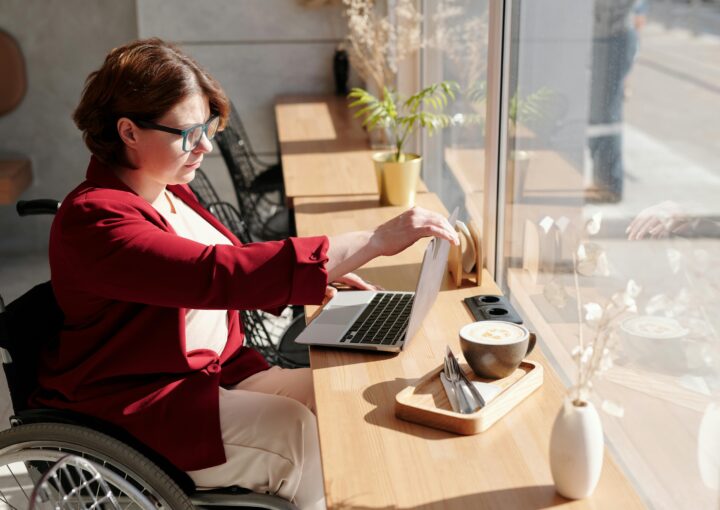 Jeune femme sur son fauteuil rulant placé devant un bureau où est placé un ordinateur portable sur lequelle elle semble travailler.